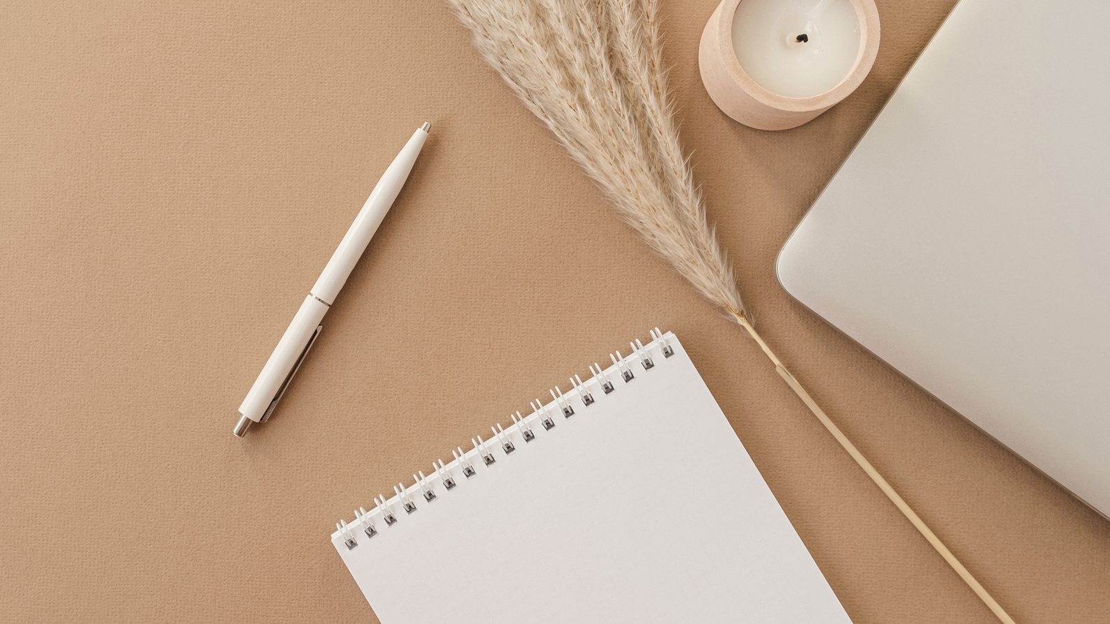 A calm desk: a blank spiral notepad, a pen, a candle and a closed laptop on a warm tan surface